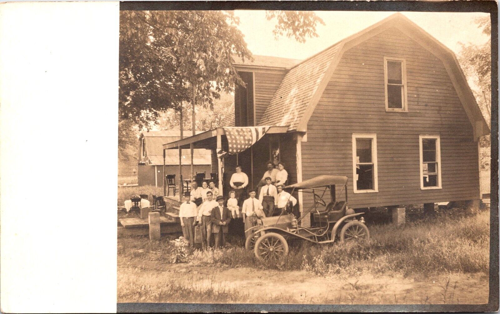 THIRTEEN PEOPLE ON FRONT PORCH OF HOUSE :AMERICAN FLAG :OLD CAR : RPPC 1904-1918