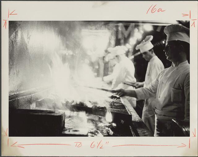 Photo:[[Line cooks in restaurant cooking in kitchen over stove]]